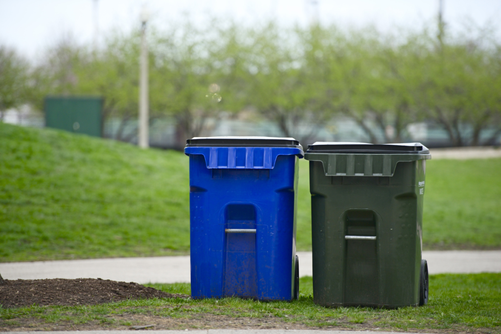 Blue and black recycling bins outside on a grassy area beside a sidewalk.