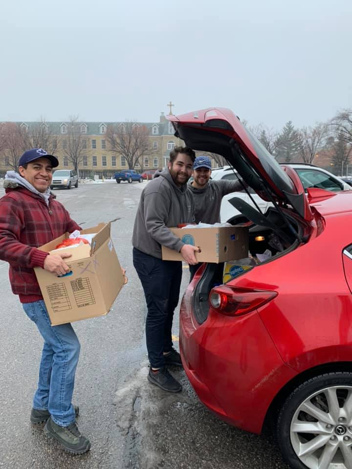 Three men smiling while unloading Christmas hampers from the trunk of a red car in a snowy parking lot.
