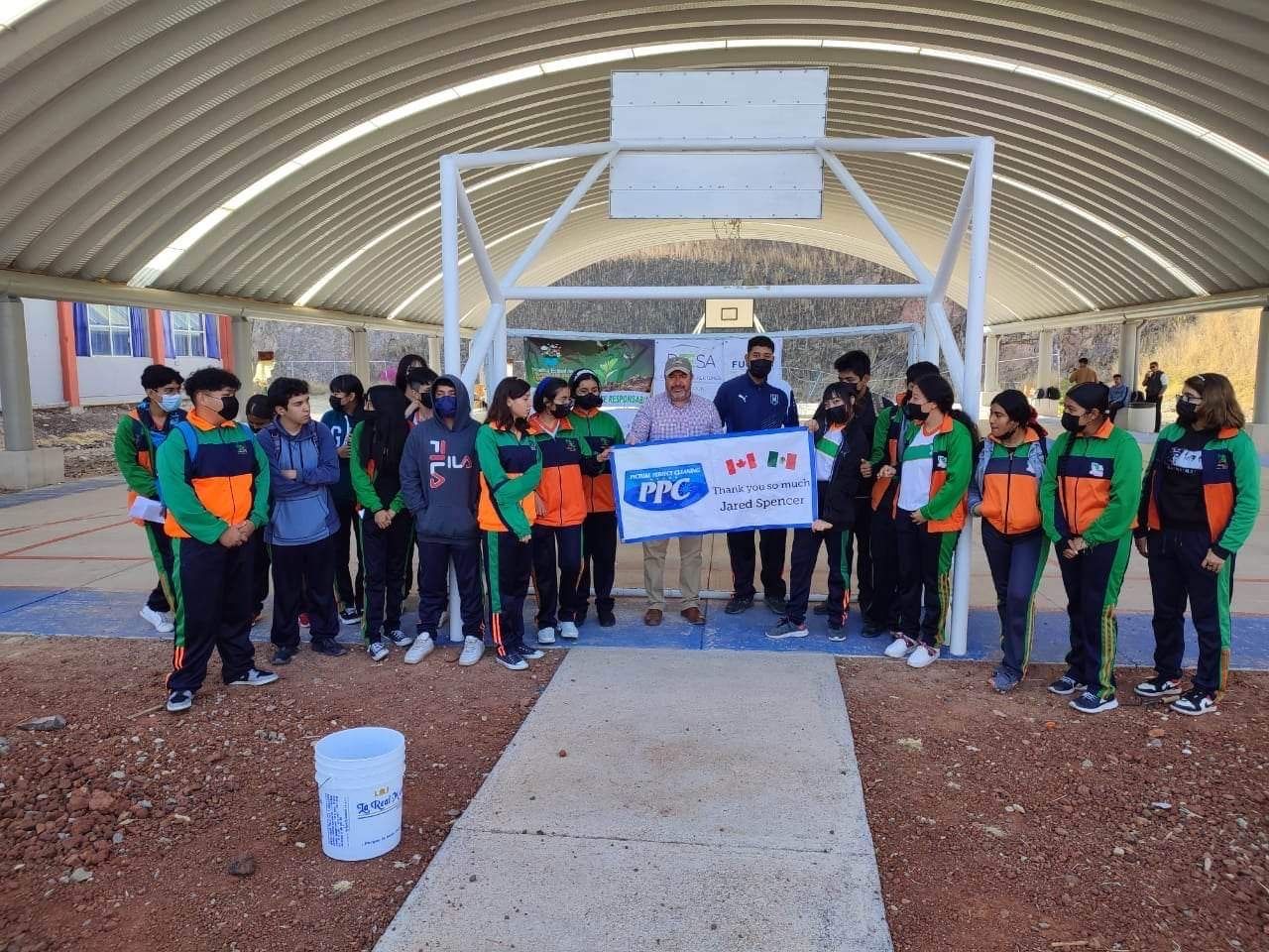 Group of Mexican students in green and orange uniforms standing with PPC team members under a covered basketball court holding a banner thanking Jared Sarbit and Picture Perfect Cleaning.
